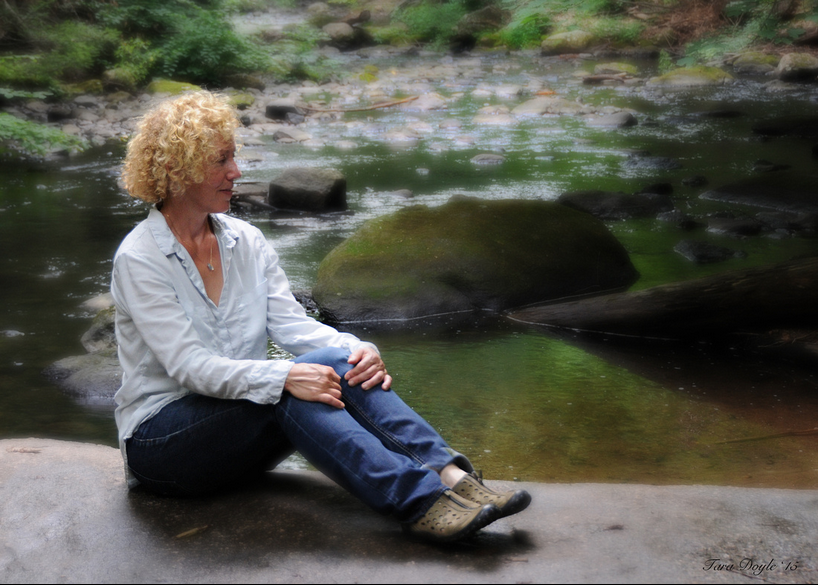 Author, Susanne Davis, sitting on a flat rock beside a green river. She has curly blond hair, and wears comfortable hiker's clothing.